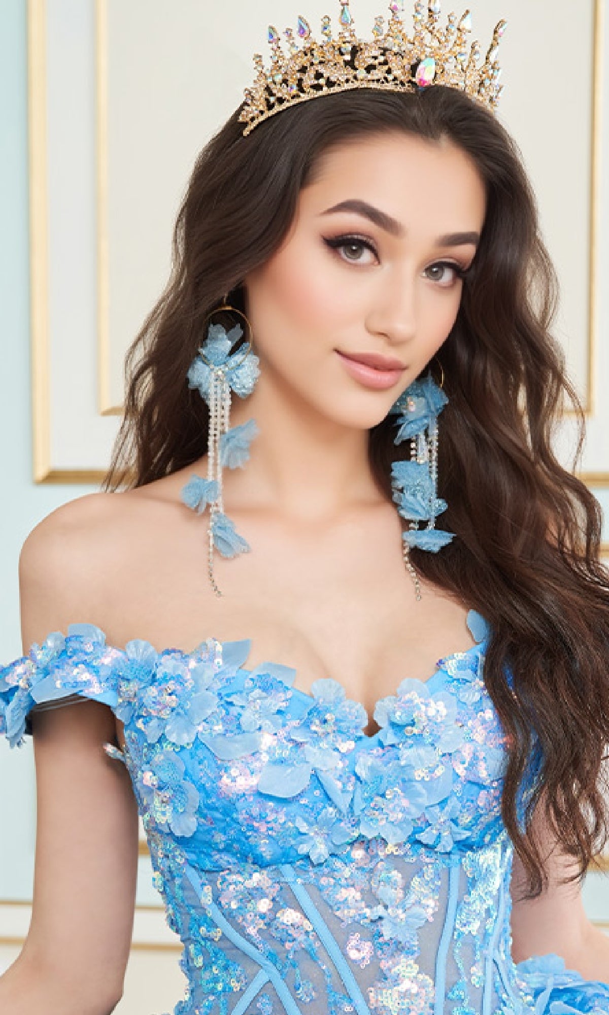 A woman wearing the Quinceanera Dress PR30162 By Princesa, featuring a jeweled crown, large dangling earrings, and an off-the-shoulder blue floral glitter tulle gown, stands before a light-colored background.